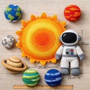 May include: A collection of crocheted space-themed toys, including a sun, planets, and an astronaut. The astronaut is 20 cm tall and the sun is 20 cm in diameter. The planets are various colors, and the sun is yellow and orange.