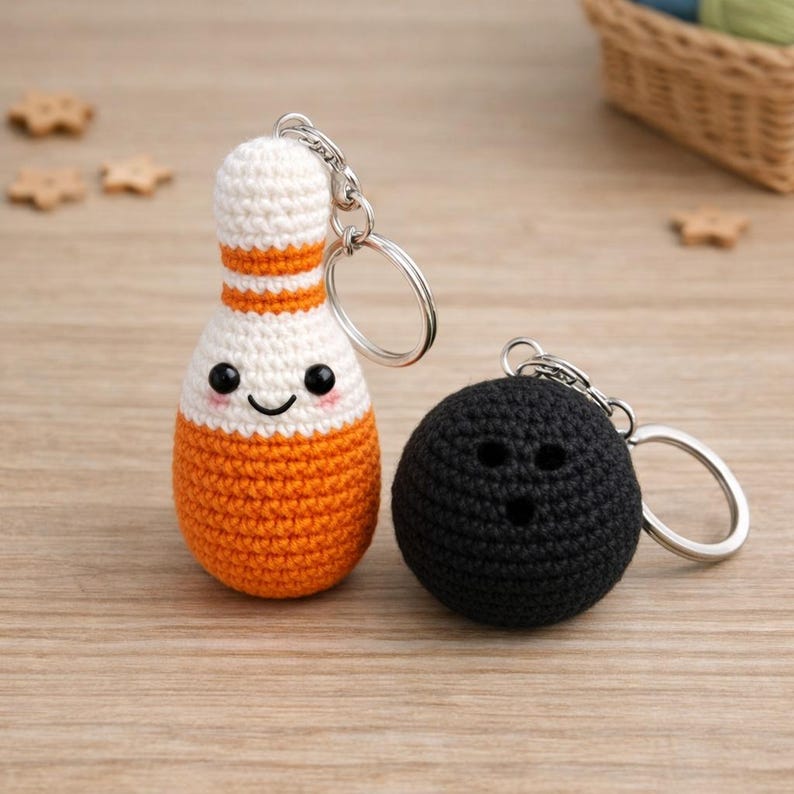 Puede incluir: Dos llaveros de ganchillo hechos a mano: un bolo y una bola de bolos. El bolo es blanco y naranja con una cara sonriente. La bola de bolos es negra con tres agujeros. Ambos tienen llaveros plateados.