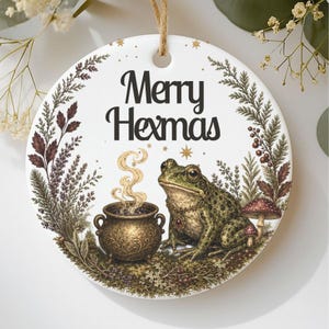 May include: A round white ceramic ornament featuring the black text Merry Hexmas. The design includes a green frog wearing a necklace, a steaming golden cauldron, and surrounding botanical elements like sprigs, berries, and mushrooms.