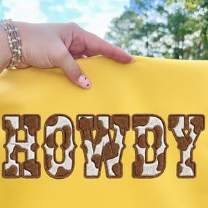 Puede incluir: Una tela amarilla con la palabra "HOWDY" bordada en letras marrones y blancas con estampado de vaca. La tela está sujeta por una mano que lleva una pulsera. El fondo está borroso con árboles verdes y cielo azul.