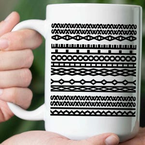 Peut inclure: Mug en céramique blanche avec un motif géométrique noir. Le mug présente diverses bandes horizontales de formes et de motifs répétitifs. Le mug est tenu dans les mains d'une personne.