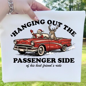 May include: White tote bag featuring a red vintage car with Santa Claus and a reindeer. The text reads "Hanging Out The Passenger Side of his best friend's ride."