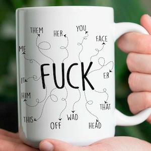 May include: White ceramic mug with the word "FUCK" in large black letters, surrounded by smaller words and arrows. The mug is held by a person. The mug has a white handle.