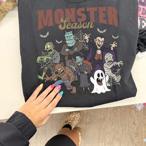 Könnte beinhalten: Ein schwarzes T-Shirt mit dem Text Monster Season in roter und goldener Schrift. Die Grafik zeigt eine Gruppe von Cartoon-Monstern, darunter ein Zombie, Frankensteins Monster, ein Werwolf, eine Hexe, ein Vampir, eine Mumie und ein Geist.