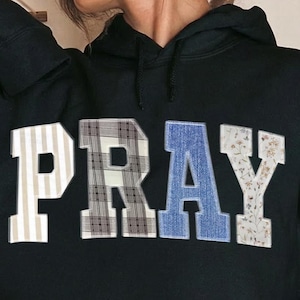Könnte beinhalten: Schwarzer Kapuzenpullover mit dem Wort "PRAY" in einem Patchwork-Design. Die Buchstaben sind weiß, blau und gemustert, was einen einzigartigen Look erzeugt. Der Pullover hat eine Vordertasche und eine Kapuze mit Kordelzug.