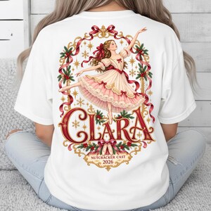 Op de afbeelding: Een wit t-shirt met een achteropdruk van een ballerina in een crème en roze jurk, omlijst door rode linten, gouden filigraan en hulst. De tekst luidt CLARA, NUTCRACKER CAST en 2026, met balletschoenen afgebeeld in het midden onderaan.