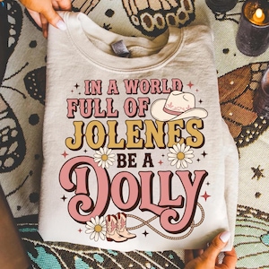 Può includere: Una felpa girocollo beige con una stampa grafica che riporta il testo IN A WORLD FULL OF JOLENES BE A DOLLY. Il design include un cappello da cowboy, due paia di stivali da cowboy, margherite e stelle nei toni del rosa, giallo e marrone.