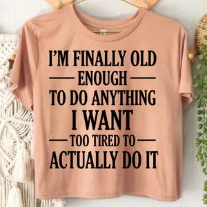 May include: A peach-coloured short-sleeve t-shirt featuring black text that reads: I'M FINALLY OLD ENOUGH TO DO ANYTHING I WANT TOO TIRED TO ACTUALLY DO IT. The shirt is displayed on a gold-toned hanger, and two red circles with white text read SVG and PNG.