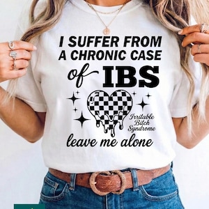 Puede incluir: Camiseta blanca con el texto negro "I SUFFER FROM A CHRONIC CASE of IBS". Un corazón a cuadros con bordes goteando, las palabras "Irritable Bitch Syndrome" y "leave me alone" completan el diseño.