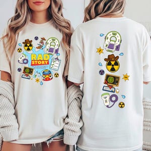 May include: A cream-colored t-shirt featuring a graphic print of medical imaging equipment, a cowboy hat, a radiation symbol, and the text "RAD STORY TO INFINITY AND BEYOND OUR PATIENTS". The back of the shirt displays a vertical arrangement of these icons.
