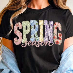 Könnte beinhalten: Schwarzes T-Shirt mit dem Wort "SPRING" in großen, farbenfrohen Buchstaben mit Blumenmuster. Das Wort "season" ist in einer Schreibschrift unter "SPRING" geschrieben. Das Shirt ist aus einem weichen Material.