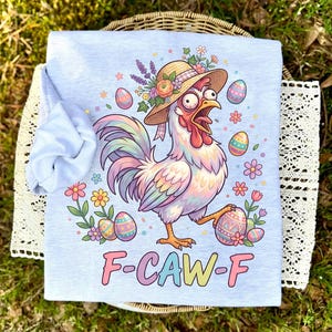 May include: A light blue t-shirt featuring a cartoon rooster wearing a hat and holding an Easter egg. The rooster is surrounded by colorful Easter eggs and flowers. The text "F-CAW-F" is printed on the shirt.