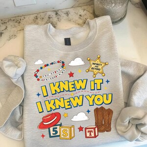 May include: A light gray crewneck sweatshirt featuring a graphic print with a sheriff badge labeled Infinity and Beyond, a red cowboy hat, brown cowboy boots, alphabet blocks, and a beaded bracelet with the text You've Got A Friend In Me.