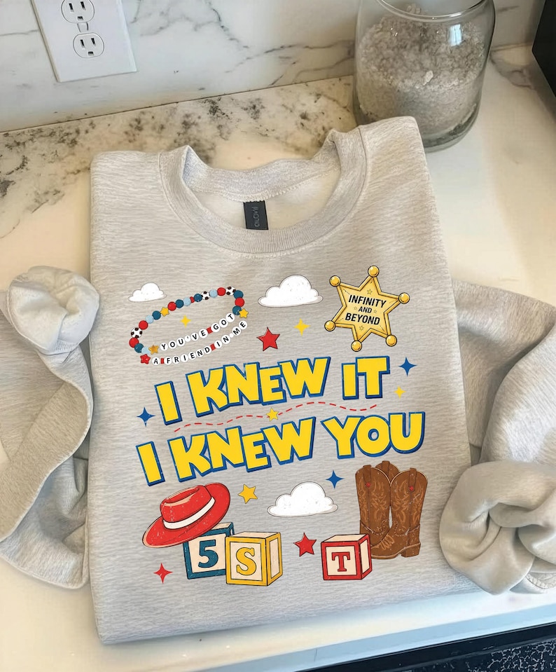 May include: A light gray crewneck sweatshirt featuring a graphic print with a sheriff badge labeled Infinity and Beyond, a red cowboy hat, brown cowboy boots, alphabet blocks, and a beaded bracelet with the text You've Got A Friend In Me.