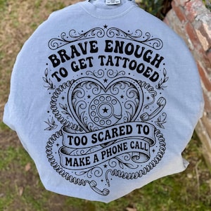 May include: A light blue sweatshirt with a black graphic design. The design features the text "Brave Enough to Get Tattooed, Too Scared to Make a Phone Call" around a heart-shaped telephone dial. The sweatshirt is a comfortable, casual style.