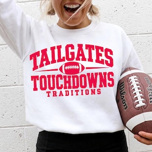 以下が含まれることがあります： TAILGATES TOUCHDOWNS TRADITIONSという赤い太字のテキストと中央にフットボールのグラフィックが描かれた、白い長袖のクルーネックトレーナー。トレーナーには、白い靴紐が付いた茶色のレザー製フットボールが添えられています。