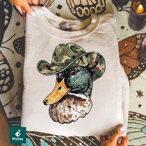 Könnte beinhalten: Beiges Sweatshirt mit detaillierter Grafik einer Ente mit Tarn-Cowboyhut. Der Entenkopf ist grün, braun und goldfarben, mit detaillierter Perlenstruktur. Das Sweatshirt ist gefaltet, wobei die Grafik gut sichtbar ist.