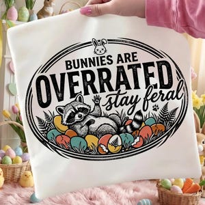 May include: White graphic tee featuring a raccoon lounging among colorful Easter eggs. The text reads "Bunnies Are Overrated Stay Feral" in a black oval design with a bunny illustration. A paw print is also included.
