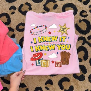 May include: A light pink t-shirt featuring a graphic print with the text "I KNEW IT I KNEW YOU" in yellow, a red cowboy hat, brown cowboy boots, a sheriff badge with "INFINITY AND BEYOND", alphabet blocks, clouds, and a beaded bracelet.