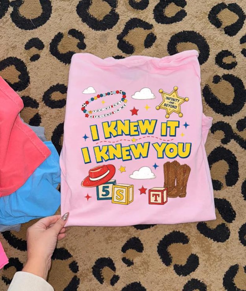 May include: A light pink t-shirt featuring a graphic print with the text "I KNEW IT I KNEW YOU" in yellow, a red cowboy hat, brown cowboy boots, a sheriff badge with "INFINITY AND BEYOND", alphabet blocks, clouds, and a beaded bracelet.