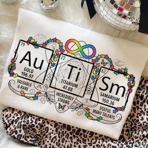 May include: White pillow with a graphic design featuring the elements Au (Gold), Ti (Titanium), and Sm (Samarium) in a periodic table style. The design includes the infinity symbol, puzzle pieces, and the text "Valuable & Rare", "Incredibly Strong", and "Useful Many Talents".