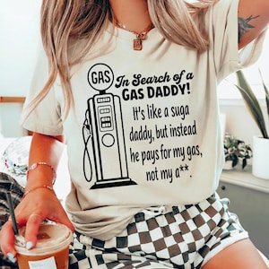 May include: Cream-colored t-shirt with a black graphic of a vintage gas pump and the text "In Search of a GAS DADDY!" The shirt's message continues with "It's like a suga daddy, but instead he pays for my gas, not my a**."