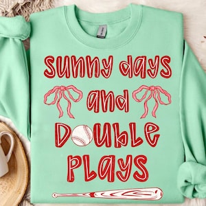 May include: A pale green sweatshirt featuring the text "SUNNY DAYS and DOUBLE PLAYS" in red. The design includes a baseball, two pink bows, and a baseball bat.