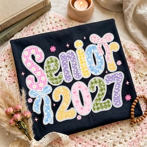 以下が含まれることがあります： 黒いTシャツに、大きくカラフルな柄入りの文字で「Senior 2027」とプリントされています。各文字は白いレースで縁取られ、ピンク、黄色、青、緑の色合いの花柄、ギンガムチェック、リボンのモチーフで装飾されています。