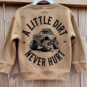 May include: A tan-coloured sweatshirt featuring a black monster truck graphic and the text "A LITTLE DIRT NEVER HURT". The crew neck sweatshirt has long sleeves. The design includes lightning bolt details.