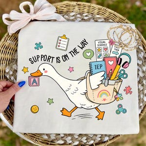 Könnte beinhalten: Ein weißes T-Shirt mit einer Cartoon-Gans, die eine Tasche voller Schulsachen trägt, darunter ein mit IEP beschriftetes Notizbuch, ein visueller Stundenplan, Kopfhörer und ein Bleistift. Der Text lautet SUPPORT IS ON THE WAY, umgeben von Puzzleteilen und Sternen.