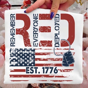 K&ouml;nnte beinhalten: Wei&szlig;es T-Shirt mit einem verblassten Design der amerikanischen Flagge. Das Shirt zeigt die Worte "REMEMBER EVERYONE DEPLOYED" in Rot und Blau, zusammen mit "EST. 1776" und einem Paar Erkennungsmarken. Das Design ist patriotisch.