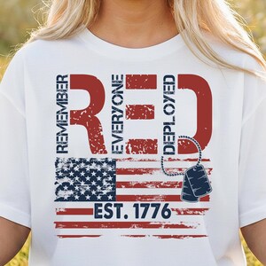 K&ouml;nnte beinhalten: Wei&szlig;es T-Shirt mit patriotischem Design. Das Design zeigt den Text "REMEMBER RED EVERYONE DEPLOYED" in Rot, zusammen mit einer amerikanischen Flagge und Erkennungsmarken. Der Text "EST. 1776" ist ebenfalls enthalten.