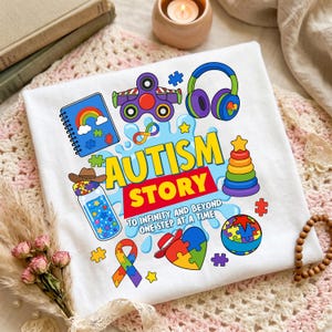 May include: A white t-shirt featuring a colorful graphic with the text "AUTISM STORY TO INFINITY AND BEYOND ONE STEP AT A TIME." The design includes a notebook, fidget spinner, headphones, a jar, a stacking toy, a ball, and puzzle pieces.