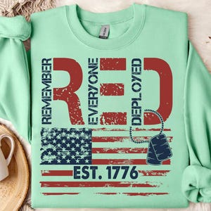 K&ouml;nnte beinhalten: Hellgr&uuml;nes Sweatshirt mit einem roten Used-Look-Design. Das Design enth&auml;lt die Worte "REMEMBER EVERYONE DEPLOYED" und eine amerikanische Flagge. Darunter der Text "EST. 1776" und Hundemarken.
