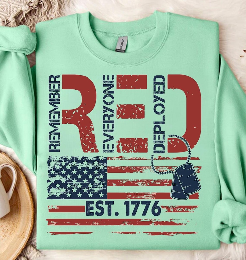 K&ouml;nnte beinhalten: Hellgr&uuml;nes Sweatshirt mit einem roten Used-Look-Design. Das Design enth&auml;lt die Worte "REMEMBER EVERYONE DEPLOYED" und eine amerikanische Flagge. Darunter der Text "EST. 1776" und Hundemarken.