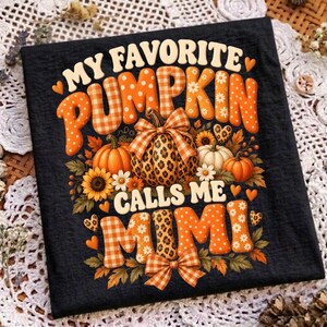 May include: A black t-shirt featuring the text MY FAVORITE PUMPKIN CALLS ME MIMI in orange and white patterned letters. The design includes illustrations of pumpkins with leopard print and plaid textures, sunflowers, leaves, and small hearts.