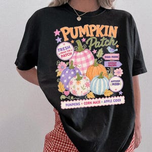 May include: A black t-shirt featuring a colourful graphic of four pumpkins in pink gingham, purple, orange, and light blue. The text reads PUMPKIN PATCH, FRESH FROM THE PATCH, FARM FRESH, LOCALLY GROWN, AUTUMN PICKED, PUMPKINS, CORN MAZE, and APPLE CIDER.