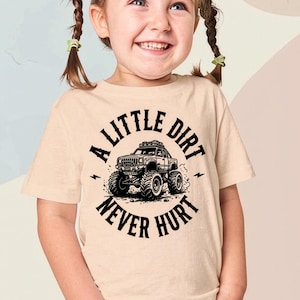 May include: A light beige t-shirt featuring a black graphic of a monster truck and the text "A LITTLE DIRT NEVER HURT". The shirt is worn by a young girl with pigtails.