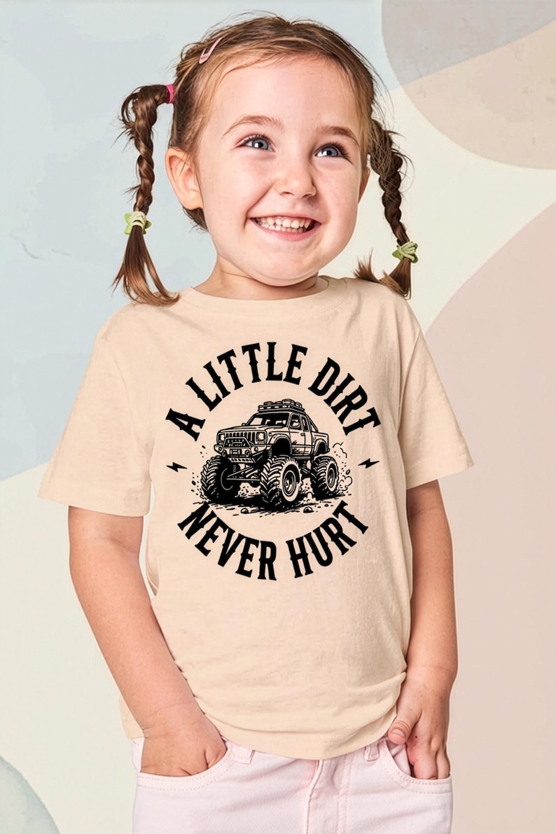 May include: A light beige t-shirt featuring a black graphic of a monster truck and the text "A LITTLE DIRT NEVER HURT". The shirt is worn by a young girl with pigtails.
