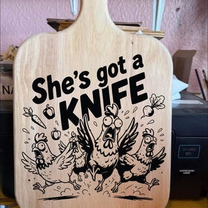 Op de afbeelding: Een houten snijplank met een handvat, met de tekst "She's got a KNIFE" in zwart. Onder de tekst staan illustraties van geschrokken kippen en groenten. De plank is gemaakt van lichtgekleurd hout.