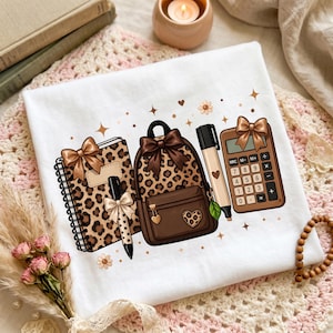Pode incluir: Uma t-shirt branca com uma impressão gráfica de um caderno de espiral com capa de leopardo, uma mochila castanha com padrão de leopardo, uma caneta preta e uma calculadora castanha, decoradas com laços castanhos e estrelas.