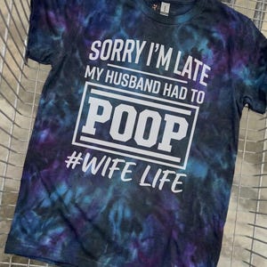 May include: A tie-dye t-shirt in shades of blue, purple, and black. The shirt features the text "SORRY I'M LATE MY HUSBAND HAD TO POOP #WIFE LIFE" in white block letters.