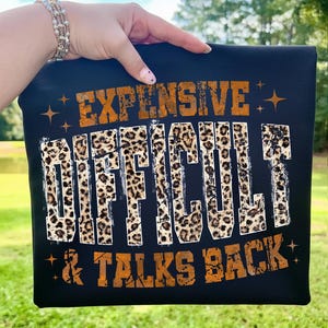May include: Black t-shirt with the words "EXPENSIVE, DIFFICULT, & TALKS BACK" in orange and leopard print lettering. The shirt is held up against a green background.