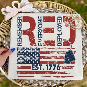 May include: Cream-colored t-shirt with a distressed American flag design. The words "REMEMBER EVERYONE DEPLOYED RED" are printed in red, along with "EST. 1776". A bow and jewelry are also visible.