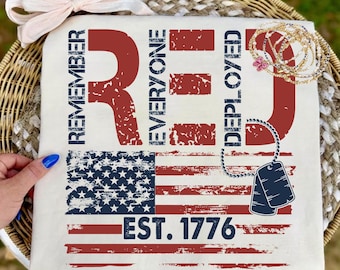 Puede incluir: Camiseta color crema con un diseño desgastado de la bandera estadounidense. Las palabras "REMEMBER EVERYONE DEPLOYED RED" están impresas en rojo, junto con "EST. 1776". También se ven un lazo y joyas.