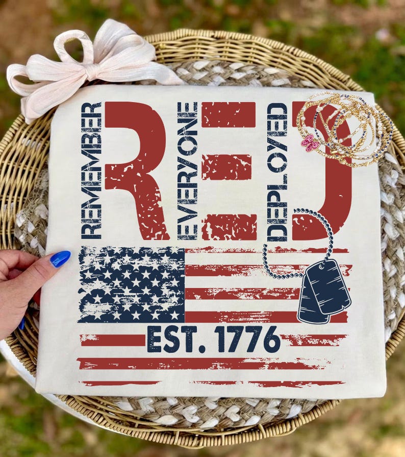 K&ouml;nnte beinhalten: Cremefarbenes T-Shirt mit einem verwitterten Design der amerikanischen Flagge. Die Worte "REMEMBER EVERYONE DEPLOYED RED" sind rot gedruckt, zusammen mit "EST. 1776". Eine Schleife und Schmuck sind ebenfalls sichtbar.
