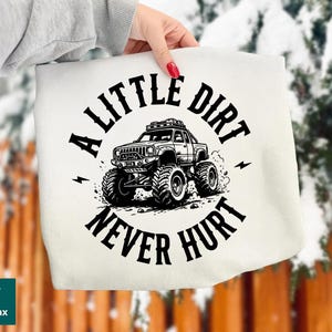 May include: A white tote bag featuring a black graphic of a monster truck with the text "A LITTLE DIRT NEVER HURT". The design includes lightning bolts and a circular layout.