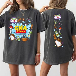 Op de afbeelding: Een antracietgrijs t-shirt met een grafische print van medische benodigdheden en cowboy-items. De voorkant toont de tekst CNA STORY TO INFINITY AND BEYOND FOR OUR PATIENTS, terwijl de achterkant een verticale rangschikking van de iconen toont.