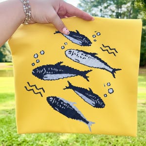 May include: Yellow pouch featuring a navy and white fish design. The fish are swimming in a sea of bubbles and wavy lines. The pouch is held by a person.