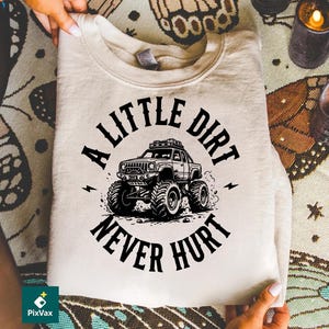 May include: A beige sweatshirt featuring a black graphic of a monster truck with the text "A Little Dirt Never Hurt." The design includes lightning bolts and a detailed illustration of the vehicle.